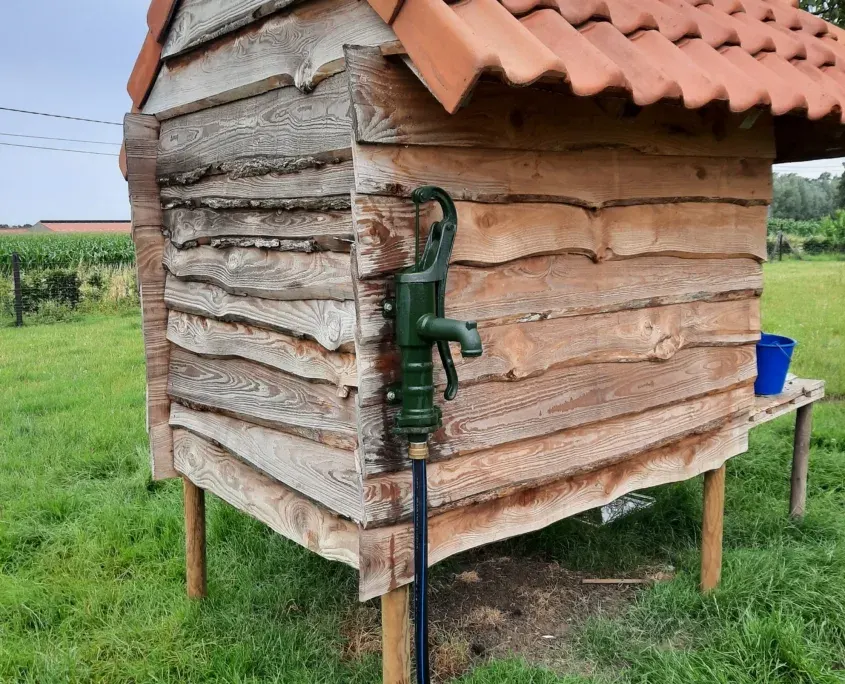 Traditionele groene gietijzeren handpomp aan een houten tuinhuisje met terracotta dak, in het gras.