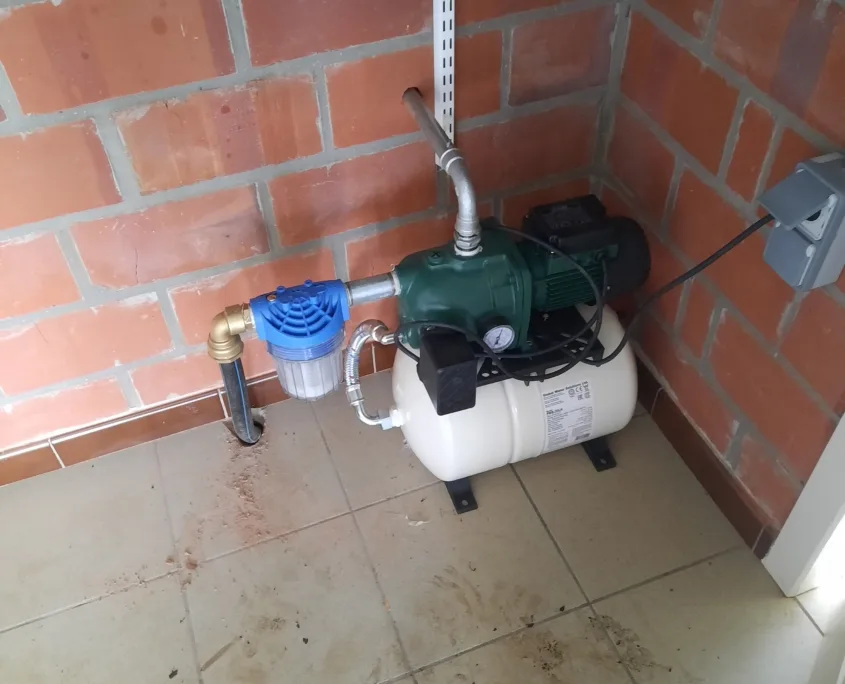 Groene elektrische centrifugaalpomp met wit hydrofoorvat en blauw voorfilter tegen een rode bakstenen muur.