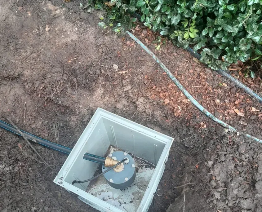 Witte kunststof pomp-inspectiekast ingegraven in de tuinaarde naast een groene haag, met zwarte kabel en slang die naar de put lopen.