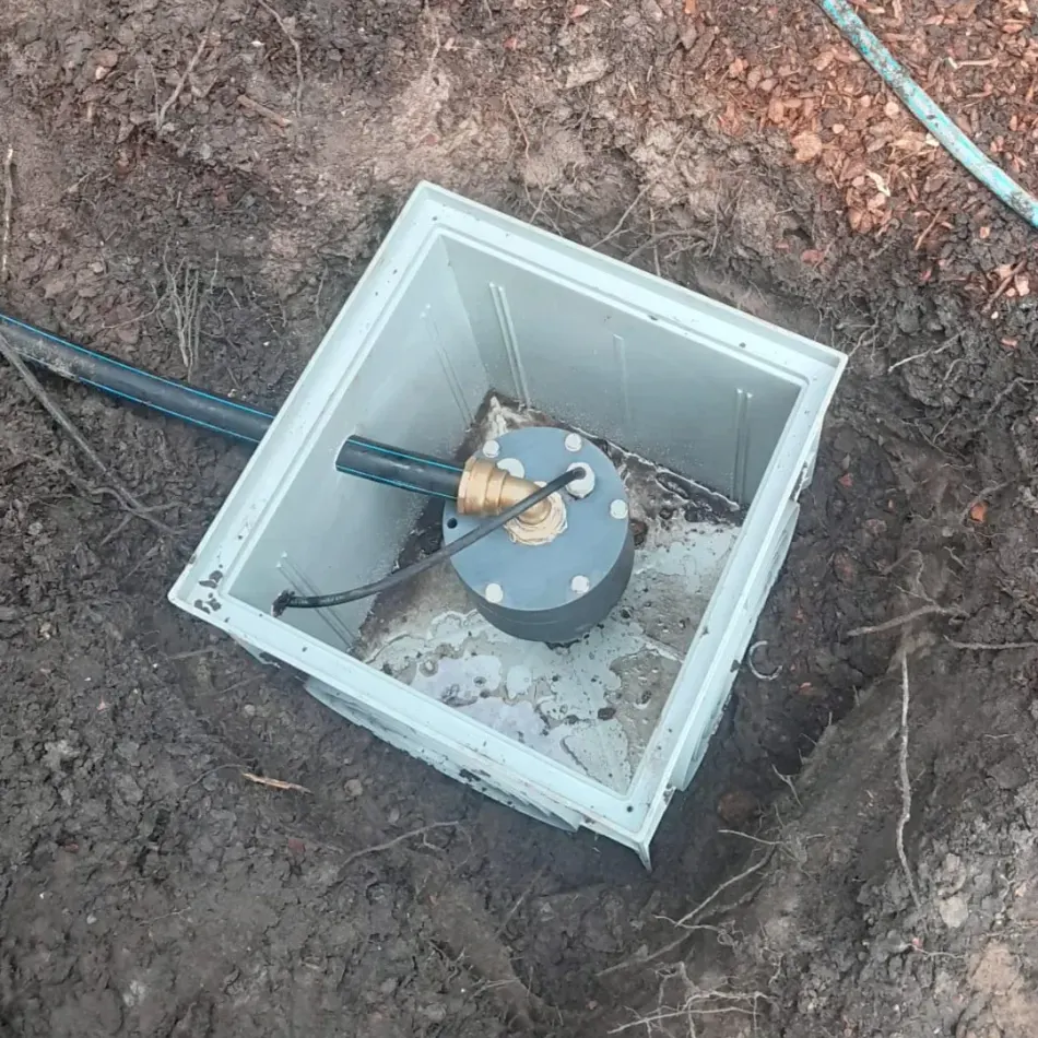 Geopende grondwaterput-inspectiekast in lichtblauw kunststof met messing pompaansluiting, ingegraven in donkere aarde.